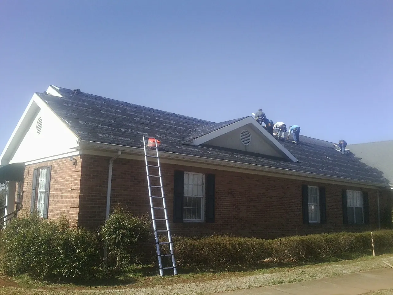 Crew tearing shingles off a brick ranch roof with a ladder set — Metal Roof Repair work by our crew in South Byron