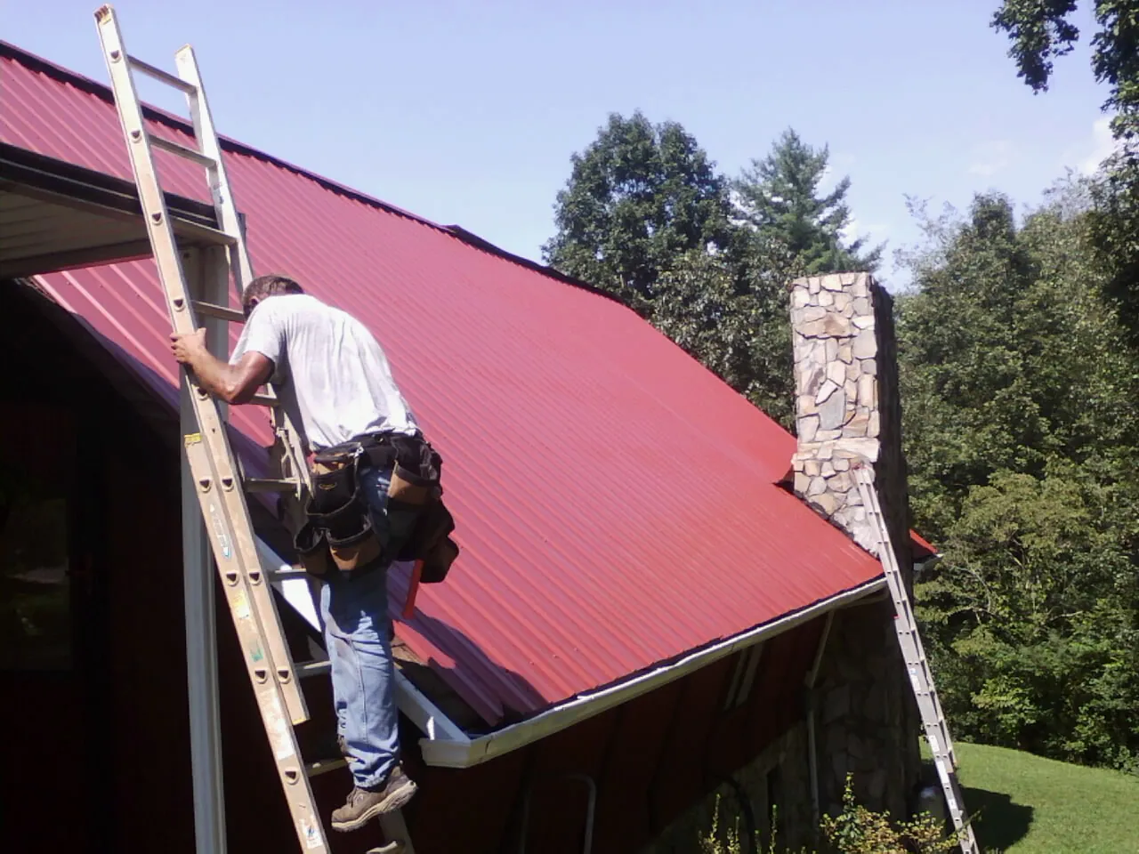 Roofer on a ladder beside a red metal roof and stone chimney — Soffit & Fascia Repair by Roofing Brothers in South Byron